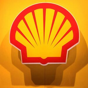 Close centralizado do logotipo da Shell, uma concha estilizada nas cores amarelo e vermelho, fixada em uma parede amarela iluminada.