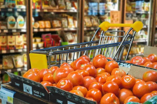 Em primeiro plano, uma caixa de papelão repleta de tomates vermelhos e maduros em um supermercado. Logo atrás da caixa, vê-se a parte superior de um carrinho de compras metálico com detalhes em amarelo. Ao fundo, as prateleiras do corredor do mercado aparecem levemente desfocadas, exibindo diversos produtos embalados. A iluminação é clara, típica de ambiente comercial, destacando a cor vibrante dos tomates.
