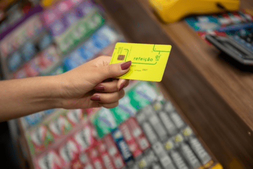 Uma pessoa segura um cartão de benefícios de cor verde-limão em frente ao caixa de um estabelecimento comercial. O cartão traz a inscrição "refeição" em letras verdes, acompanhada de pequenos ícones de talheres e de um copo com canudo. Em segundo plano, desfocados, aparecem produtos expostos perto do caixa, como maços de cigarro e gomas de mascar, além de uma máquina de cartão amarela sobre o balcão de madeira.