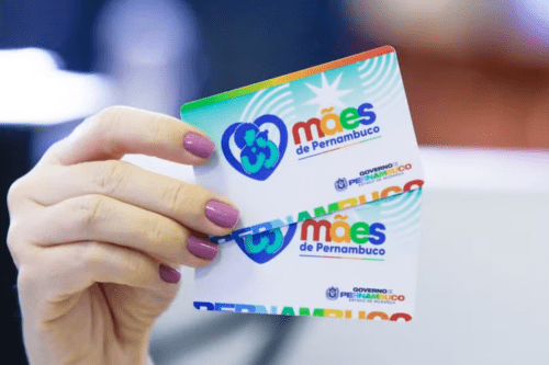 Uma mão com unhas pintadas de esmalte lilás segura dois cartões de benefício social sobrepostos. O cartão da frente está em foco e exibe, do lado esquerdo, um coração azul com a ilustração em verde de uma mãe abraçando o filho. Ao lado, está escrito o nome do programa "mães de Pernambuco", com a palavra "mães" em letras coloridas. No canto inferior direito, consta o logotipo do Governo do Estado de Pernambuco com o slogan "Estado de Mudança". O segundo cartão, idêntico, aparece logo atrás, ligeiramente deslocado. O fundo da imagem está desfocado.
