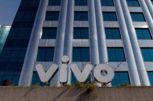 Vista externa de um edifício comercial moderno com janelas espelhadas. Na base do prédio, o logotipo prateado da operadora "Vivo" está fixado em uma mureta cinza.