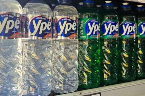Uma fileira de frascos plásticos de detergente da marca brasileira Ypê organizados em uma prateleira de supermercado. À esquerda, estão os frascos de detergente "Clear", que é totalmente transparente e límpido. À direita, estão os frascos da versão "Limão", de cor verde vibrante. As embalagens são cilíndricas, com texturas em relevo no plástico e rótulos azuis metálicos com o logotipo branco da Ypê.