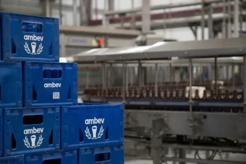 Caixas da Ambev empilhadas em fábrica com linha de produção de cerveja ao fundo.