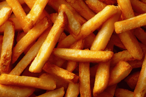 Vista aproximada e em ângulo superior de uma porção de batatas fritas douradas e crocantes, empilhadas de forma irregular. As batatas têm formato de palito grosso, com algumas bordas levemente tostadas e uma textura visivelmente frita e brilhante.