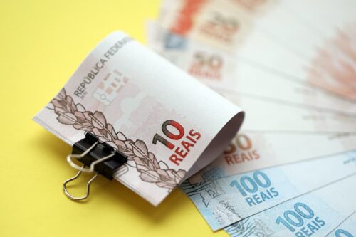 Notas de real presas com clipe, com cédulas de R$10, R$50 e R$100 ao fundo sobre superfície colorida.