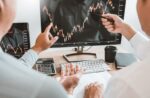Duas pessoas analisam gráfico de mercado em monitor, discutindo os melhores investimentos e tendências financeiras em ambiente de escritório.