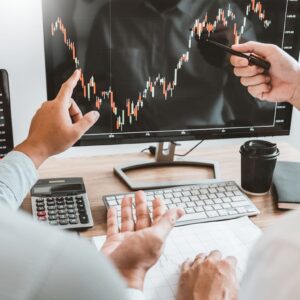 Duas pessoas analisam gráfico de mercado em monitor, discutindo os melhores investimentos e tendências financeiras em ambiente de escritório.