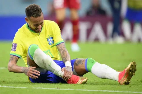 Neymar sentado no gramado com expressão de dor durante jogo da Seleção Brasileira após sofrer lesão na perna direita.