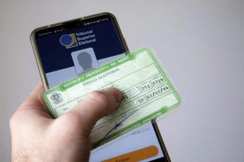 Indivíduo com um smartphone mostrando do aplicativo do Tribunal Superior Eleitoral enquanto segura um título de eleitor.