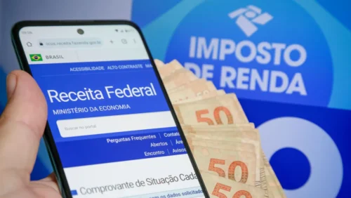 Celular acessa portal da Receita Federal ao lado de notas de real e símbolo do Imposto de Renda 2026.