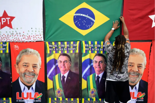 Uma parede exibe diversos cartazes e bandeiras de propaganda política brasileira. No centro superior, há uma bandeira do Brasil que está sendo ajustada por uma pessoa de costas, que possui cabelos com dreadlocks e usa uma camiseta camuflada. Ao redor da bandeira nacional, estão dispostas bandeiras brancas e vermelhas do Partido dos Trabalhadores (PT). Logo abaixo, há uma fileira de cartazes verticais intercalados: dois exibem o rosto de Luiz Inácio Lula da Silva com o slogan "Lula - O Brasil feliz de novo", e dois exibem o rosto de Jair Bolsonaro com a faixa presidencial e a frase "Brasil acima de tudo, Deus acima de todos - Fechado com Bolsonaro!".