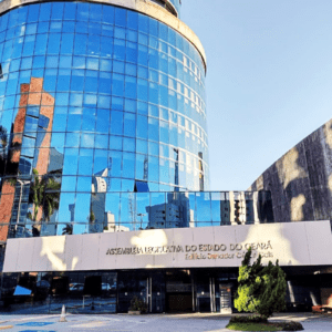 Fachada do edifício da Assembleia Legislativa do Estado do Ceará (ALECE) - Edifício Senador César Cals. A estrutura principal à esquerda é uma torre cilíndrica moderna revestida de vidro espelhado azul, que reflete o céu e coqueiros próximos. Na entrada, há uma marquise branca horizontal com o nome do órgão em letras douradas. À direita, ergue-se uma parede de concreto texturizado. O dia está ensolarado e o céu limpo.