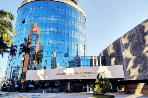 Fachada do edifício da Assembleia Legislativa do Estado do Ceará (ALECE) - Edifício Senador César Cals. A estrutura principal à esquerda é uma torre cilíndrica moderna revestida de vidro espelhado azul, que reflete o céu e coqueiros próximos. Na entrada, há uma marquise branca horizontal com o nome do órgão em letras douradas. À direita, ergue-se uma parede de concreto texturizado. O dia está ensolarado e o céu limpo.
