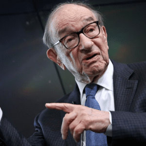 Fotografia em plano médio de Alan Greenspan, um homem idoso de pele clara e cabelos grisalhos calvo no topo. Ele usa óculos de armação grossa preta, terno risca-de-giz escuro, camisa social branca e uma gravata azul texturizada. Ele está com o corpo ligeiramente inclinado para a direita, com a mão esquerda erguida e o punho parcialmente fechado, enquanto aponta com o dedo indicador da mão direita para o lado. O fundo é escuro e desfocado, com um leve reflexo de luz colorida à direita.