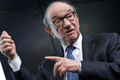 Fotografia em plano médio de Alan Greenspan, um homem idoso de pele clara e cabelos grisalhos calvo no topo. Ele usa óculos de armação grossa preta, terno risca-de-giz escuro, camisa social branca e uma gravata azul texturizada. Ele está com o corpo ligeiramente inclinado para a direita, com a mão esquerda erguida e o punho parcialmente fechado, enquanto aponta com o dedo indicador da mão direita para o lado. O fundo é escuro e desfocado, com um leve reflexo de luz colorida à direita.