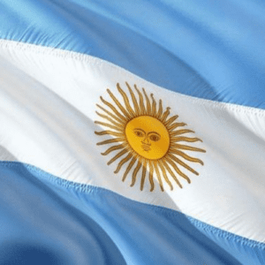 Uma imagem em plano detalhe mostra a bandeira da Argentina em uma textura de tecido ondulado. A bandeira é composta por três faixas horizontais de igual tamanho: a superior e a inferior são de cor azul-celeste, e a central é branca. No centro da faixa branca, destaca-se o Sol de Maio, um emblema dourado com um rosto humano figurativo, cercado por raios retos e ondulados que se alternam ao seu redor. O efeito de dobras no tecido cria áreas de luz e sombra, transmitindo a sensação de movimento.
