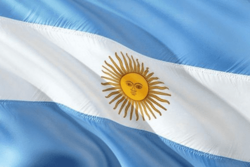 Uma imagem em plano detalhe mostra a bandeira da Argentina em uma textura de tecido ondulado. A bandeira é composta por três faixas horizontais de igual tamanho: a superior e a inferior são de cor azul-celeste, e a central é branca. No centro da faixa branca, destaca-se o Sol de Maio, um emblema dourado com um rosto humano figurativo, cercado por raios retos e ondulados que se alternam ao seu redor. O efeito de dobras no tecido cria áreas de luz e sombra, transmitindo a sensação de movimento.