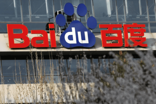 Fachada de um edifício com o logotipo da Baidu em destaque. O nome "Baidu" está escrito com as letras "Bai" e os caracteres chineses "百度" em vermelho, enquanto as letras "du" estão em branco, inseridas dentro de uma grande pegada de animal azul. Em primeiro plano, a imagem mostra galhos de árvores e flores levemente desfocadas.