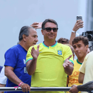 Fotografia colorida em plano médio de um grupo de homens em um evento público ao ar livre. No centro, em destaque, um homem sorri usando óculos de sol escuros e a camiseta amarela da seleção brasileira de futebol, com as mãos levantadas como se estivesse aplaudindo. À esquerda dele, um homem de camiseta azul olha para o lado. À direita, um jovem de camiseta amarela observa a cena com a língua ligeiramente para fora. Ao fundo, braços seguram celulares para registrar o momento.