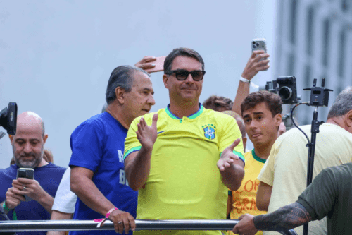 Fotografia colorida em plano médio de um grupo de homens em um evento público ao ar livre. No centro, em destaque, um homem sorri usando óculos de sol escuros e a camiseta amarela da seleção brasileira de futebol, com as mãos levantadas como se estivesse aplaudindo. À esquerda dele, um homem de camiseta azul olha para o lado. À direita, um jovem de camiseta amarela observa a cena com a língua ligeiramente para fora. Ao fundo, braços seguram celulares para registrar o momento.