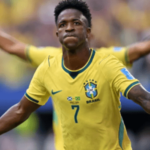 Fotografia em plano médio do jogador de futebol Vinicius Junior vestindo a camisa amarela número 7 da Seleção Brasileira. Ele aparece em primeiro plano com os braços abertos, uma expressão facial séria e lábios cerrados, parecendo comemorar em campo. No peito da camisa, além do escudo da CBF, há uma pequena estampa com as bandeiras da Escócia e do Brasil. Ao fundo, desfocado, outro jogador com os braços erguidos e a torcida em um estádio.