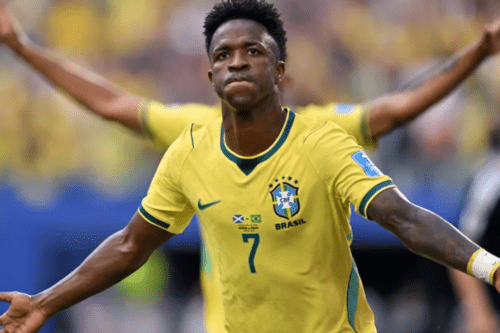 Fotografia em plano médio do jogador de futebol Vinicius Junior vestindo a camisa amarela número 7 da Seleção Brasileira. Ele aparece em primeiro plano com os braços abertos, uma expressão facial séria e lábios cerrados, parecendo comemorar em campo. No peito da camisa, além do escudo da CBF, há uma pequena estampa com as bandeiras da Escócia e do Brasil. Ao fundo, desfocado, outro jogador com os braços erguidos e a torcida em um estádio.