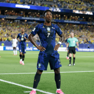 Um jogador de futebol negro com o uniforme azul-escuro da Seleção Brasileira (camisa de número 7 e calção combinando) está em pé no centro do campo, com as mãos na cintura e uma expressão facial de surpresa ou comemoração (boca aberta em formato de "O").