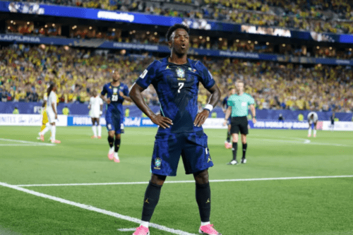 Um jogador de futebol negro com o uniforme azul-escuro da Seleção Brasileira (camisa de número 7 e calção combinando) está em pé no centro do campo, com as mãos na cintura e uma expressão facial de surpresa ou comemoração (boca aberta em formato de "O").