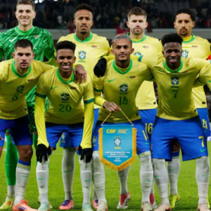Foto oficial da escalação inicial da Seleção Brasileira Masculina de Futebol em campo, posicionada em duas fileiras antes de uma partida. Todos vestem o uniforme tradicional com a camisa amarela e calções azuis, exceto o goleiro, que está em pé na fileira de trás vestindo um uniforme totalmente verde. Os jogadores estão abraçados pelos ombros. Na fileira da frente, no centro, um jogador segura uma flâmula azul da CBF (Confederação Brasileira de Futebol). O fundo mostra um estádio iluminado com torcedores nas arquibancadas.
