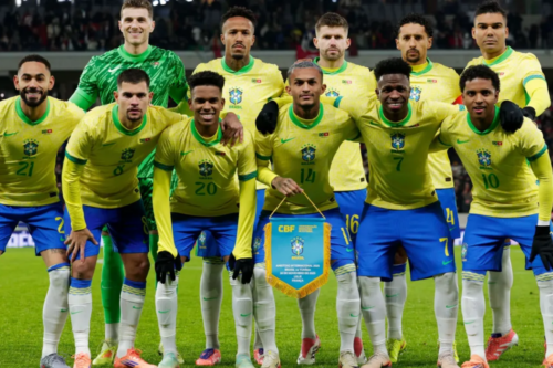Foto oficial da escalação inicial da Seleção Brasileira Masculina de Futebol em campo, posicionada em duas fileiras antes de uma partida. Todos vestem o uniforme tradicional com a camisa amarela e calções azuis, exceto o goleiro, que está em pé na fileira de trás vestindo um uniforme totalmente verde. Os jogadores estão abraçados pelos ombros. Na fileira da frente, no centro, um jogador segura uma flâmula azul da CBF (Confederação Brasileira de Futebol). O fundo mostra um estádio iluminado com torcedores nas arquibancadas.