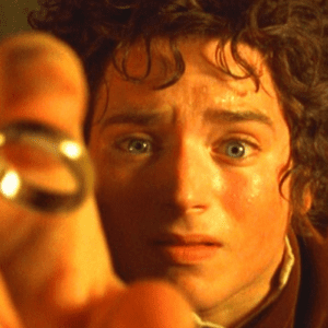 Close-up do personagem Frodo Bolseiro, interpretado por Elijah Wood nos filmes de "O Senhor dos Anéis". Ele tem cabelos escuros e cacheados, olhos azuis bem abertos com uma expressão de fascínio e apreensão, e o rosto levemente suado, iluminado por uma luz quente e amarelada. Em primeiro plano, desfocada, a mão dele segura o Um Anel em direção à câmera.