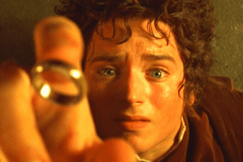 Close-up do personagem Frodo Bolseiro, interpretado por Elijah Wood nos filmes de "O Senhor dos Anéis". Ele tem cabelos escuros e cacheados, olhos azuis bem abertos com uma expressão de fascínio e apreensão, e o rosto levemente suado, iluminado por uma luz quente e amarelada. Em primeiro plano, desfocada, a mão dele segura o Um Anel em direção à câmera.