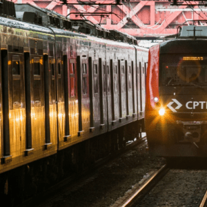 Dois trens vermelhos da CPTM (Companhia Paulista de Trens Metropolitanos) em trilhos paralelos. O trem da esquerda está de perfil, com reflexos de luz em suas janelas e portas. O trem da direita está de frente, com os faróis acesos iluminando os trilhos e o logotipo branco da CPTM destacado na parte frontal. Ao fundo, uma estrutura metálica vermelha de uma ponte ou passarela cruza acima dos trilhos.