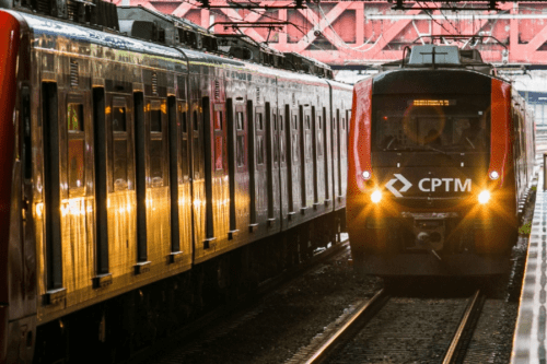 Dois trens vermelhos da CPTM (Companhia Paulista de Trens Metropolitanos) em trilhos paralelos. O trem da esquerda está de perfil, com reflexos de luz em suas janelas e portas. O trem da direita está de frente, com os faróis acesos iluminando os trilhos e o logotipo branco da CPTM destacado na parte frontal. Ao fundo, uma estrutura metálica vermelha de uma ponte ou passarela cruza acima dos trilhos.