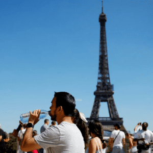 Turistas em frente à Torre Eiffel, em Paris, tomando água e tirando fotos com o céu azul ao fundo. Pessoas visitando o monumento no período diurno.