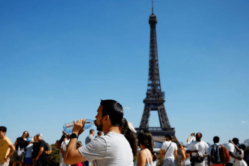 Turistas em frente à Torre Eiffel, em Paris, tomando água e tirando fotos com o céu azul ao fundo. Pessoas visitando o monumento no período diurno.
