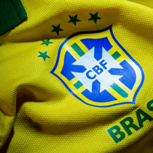 Foto em plano detalhe do escudo da Seleção Brasileira de Futebol em uma camisa amarela oficial. O escudo da CBF (Confederação Brasileira de Futebol) está centralizado, em formato de escudo azul e branco com uma cruz estilizada verde e amarela no centro. Acima do escudo, há cinco estrelas verdes dispostas em arco, representando o pentacampeonato mundial. Abaixo, está escrita a palavra "BRASIL" em letras maiúsculas verdes. O tecido da camisa é amarelo vibrante e possui uma textura canelada com pequenos furos respiráveis, mostrando dobras e sombras suaves. No canto superior esquerdo, aparece um detalhe da gola verde.