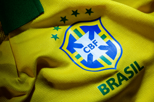 Foto em plano detalhe do escudo da Seleção Brasileira de Futebol em uma camisa amarela oficial. O escudo da CBF (Confederação Brasileira de Futebol) está centralizado, em formato de escudo azul e branco com uma cruz estilizada verde e amarela no centro. Acima do escudo, há cinco estrelas verdes dispostas em arco, representando o pentacampeonato mundial. Abaixo, está escrita a palavra "BRASIL" em letras maiúsculas verdes. O tecido da camisa é amarelo vibrante e possui uma textura canelada com pequenos furos respiráveis, mostrando dobras e sombras suaves. No canto superior esquerdo, aparece um detalhe da gola verde.