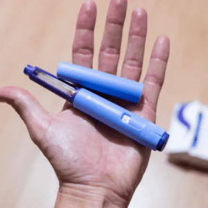 Uma foto em plano detalhe mostra a palma de uma mão aberta segurando uma caneta injetora de medicamento na cor azul claro, com a tampa removida e posicionada ao lado. Ao fundo, fora de foco, há uma superfície de madeira clara e parte de uma caixa de remédio branca com detalhes em azul.