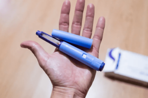 Uma foto em plano detalhe mostra a palma de uma mão aberta segurando uma caneta injetora de medicamento na cor azul claro, com a tampa removida e posicionada ao lado. Ao fundo, fora de foco, há uma superfície de madeira clara e parte de uma caixa de remédio branca com detalhes em azul.