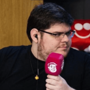 O streamer e jornalista Casimiro Miguel (Cazé) de perfil, usando camiseta preta e óculos, enquanto segura um microfone rosa com o logotipo de seu canal.