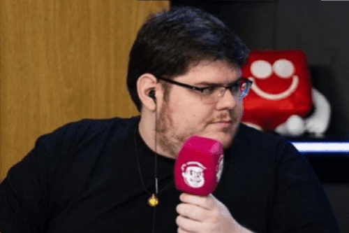O streamer e jornalista Casimiro Miguel (Cazé) de perfil, usando camiseta preta e óculos, enquanto segura um microfone rosa com o logotipo de seu canal.