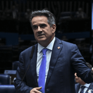 Uma imagem em plano médio mostra um homem de pele clara e cabelos escuros levemente grisalhos discursando em um plenário político. Ele veste um terno azul-marinho, camisa branca e uma gravata azul-clara. O homem está posicionado do lado esquerdo do centro, voltado para a direita, com a boca aberta como se falasse ao microfone fixado à sua frente. Sua mão esquerda está estendida para o lado com os dedos abertos em um gesto expressivo. Ao fundo, as fileiras de poltronas azuis do plenário estão desfocadas, e parte de outra pessoa de terno é visível no canto inferior direito.