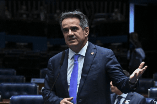 Uma imagem em plano médio mostra um homem de pele clara e cabelos escuros levemente grisalhos discursando em um plenário político. Ele veste um terno azul-marinho, camisa branca e uma gravata azul-clara. O homem está posicionado do lado esquerdo do centro, voltado para a direita, com a boca aberta como se falasse ao microfone fixado à sua frente. Sua mão esquerda está estendida para o lado com os dedos abertos em um gesto expressivo. Ao fundo, as fileiras de poltronas azuis do plenário estão desfocadas, e parte de outra pessoa de terno é visível no canto inferior direito.