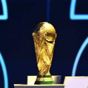 Fotografia em primeiro plano do troféu dourado da Copa do Mundo da FIFA, posicionado sobre um pedestal. A escultura detalhada representa duas figuras humanas estilizadas segurando o planeta Terra. O fundo está desfocado em tons de azul escuro com linhas curvas luminosas na cor azul claro.