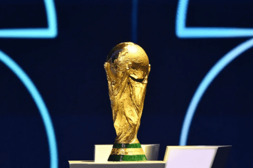 Fotografia em primeiro plano do troféu dourado da Copa do Mundo da FIFA, posicionado sobre um pedestal. A escultura detalhada representa duas figuras humanas estilizadas segurando o planeta Terra. O fundo está desfocado em tons de azul escuro com linhas curvas luminosas na cor azul claro.