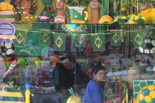 Vista através da vitrine de uma loja decorada com as cores do Brasil. Na parte superior, há chapéus, perucas e bonés verdes e amarelos em manequins, além de pequenas bandeiras do Brasil penduradas em um varal. No interior, prateleiras exibem diversos produtos, como copos e tigelas coloridas. Duas pessoas estão dentro do estabelecimento: um homem ao fundo, levemente inclinado, e uma mulher em primeiro plano à direita, olhando em direção à câmera enquanto segura um objeto longo e fino. O reflexo no vidro da vitrine mostra vislumbres da rua externa.
