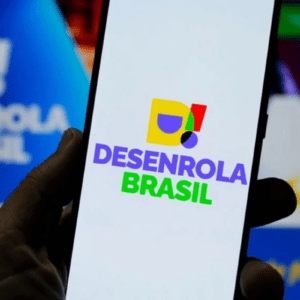 Uma pessoa segura um smartphone em primeiro plano, exibindo na tela o logotipo colorido do programa "Desenrola Brasil" sobre um fundo branco. Ao fundo, fora de foco, há uma tela de computador azul que também mostra a marca do programa.