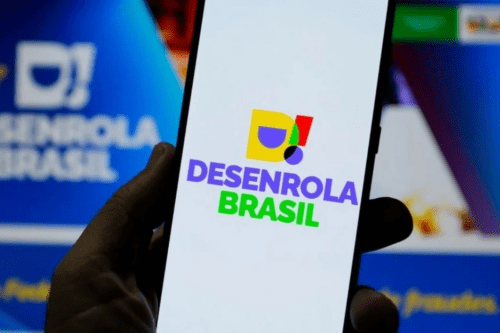 Uma pessoa segura um smartphone em primeiro plano, exibindo na tela o logotipo colorido do programa "Desenrola Brasil" sobre um fundo branco. Ao fundo, fora de foco, há uma tela de computador azul que também mostra a marca do programa.
