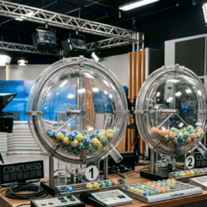 Vista em plano médio do estúdio de gravação de um sorteio de loteria, focado em dois grandes globos acrílicos transparentes e esféricos posicionados sobre uma mesa de madeira. O globo da esquerda, identificado com o número um, está repleto de bolinhas numeradas nas cores azul, amarelo e verde.